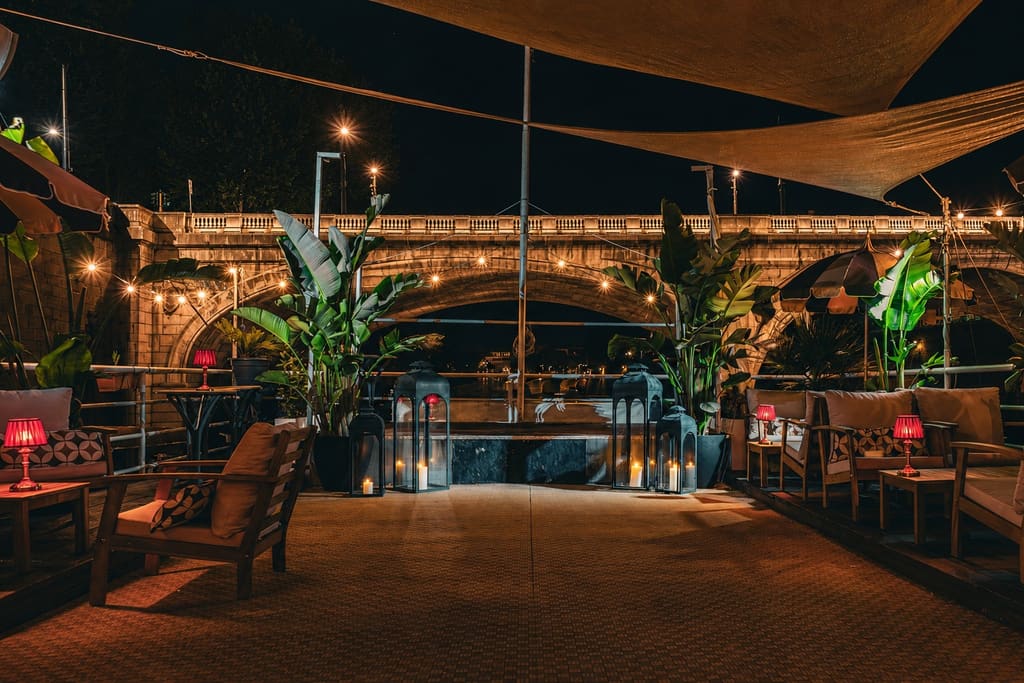 La terrazza sul Tevere di sera, con lanterne, piante, tende a vela e il ponte illuminato